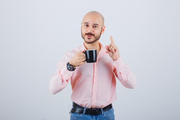 Jeune homme tenant une tasse tout en pointant vers le haut en chemise rose, jeans, vue de face.