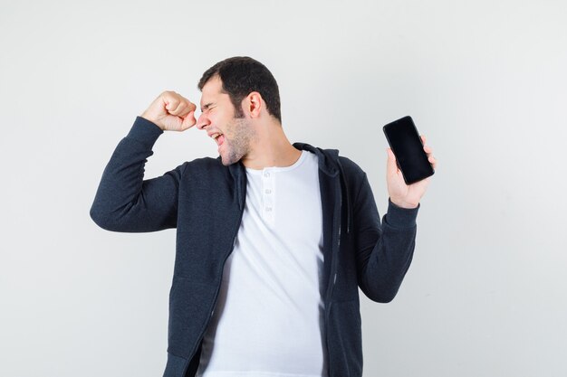 Jeune homme tenant un téléphone mobile en t-shirt, veste et air heureux. vue de face.