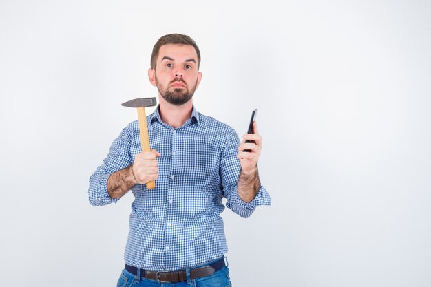 Jeune homme tenant un téléphone mobile tout en gardant le marteau en chemise, jeans et à la recherche de sérieux vue de face.