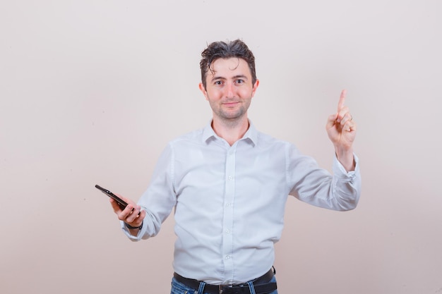 Jeune homme tenant un téléphone portable, pointant vers le haut en chemise, jeans et semblant positif