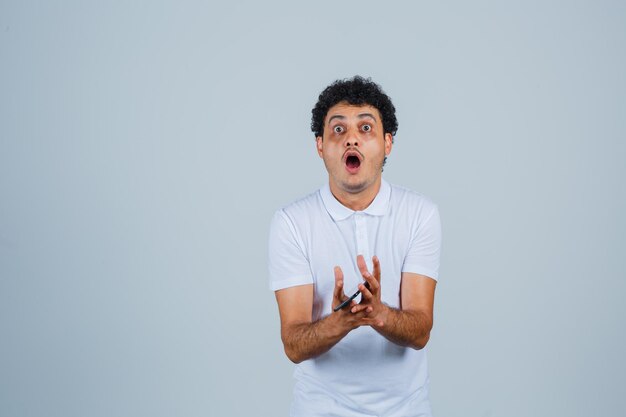 Jeune homme tenant un téléphone portable en t-shirt blanc et l'air étonné, vue de face.