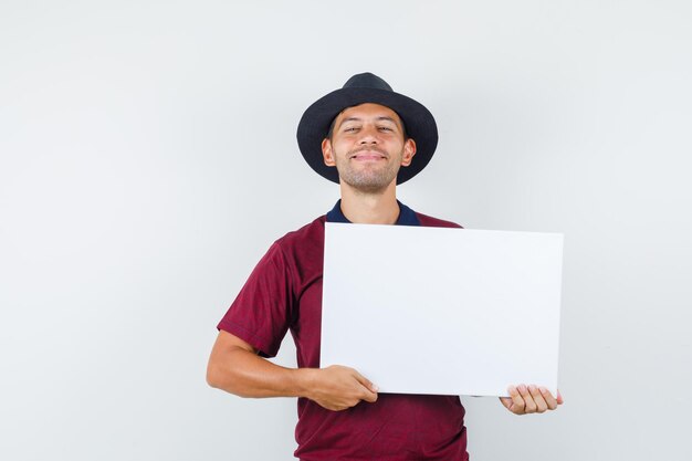 Jeune homme tenant une toile vierge en t-shirt, chapeau et l'air joyeux, vue de face.