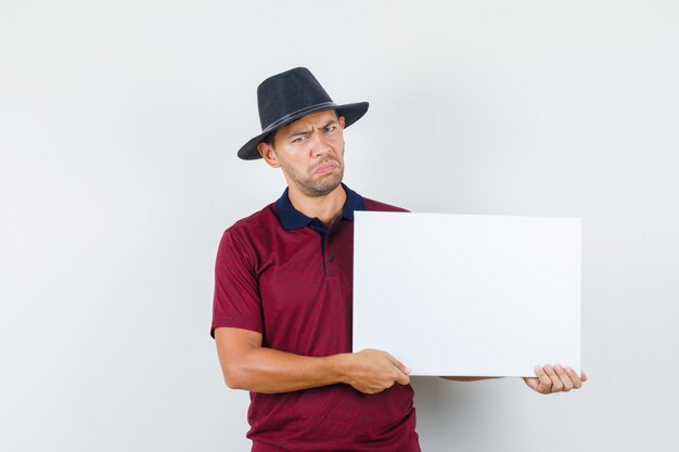 Jeune homme tenant une toile vierge en t-shirt, chapeau et à la sombre. vue de face.