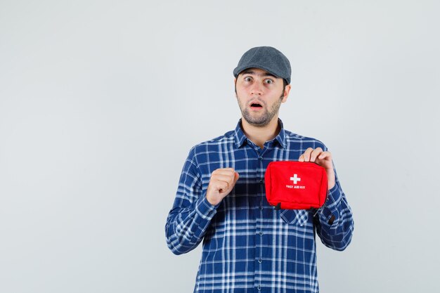 Jeune homme tenant une trousse de premiers soins en chemise, casquette et à la surprise, vue de face.