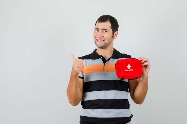 Jeune homme tenant une trousse de premiers soins tout en montrant le pouce vers le haut en t-shirt et à la satisfaction