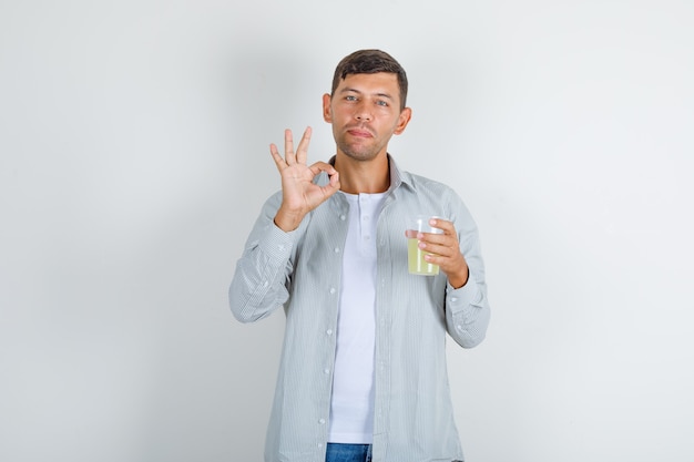Jeune homme tenant un verre de jus avec signe ok en chemise