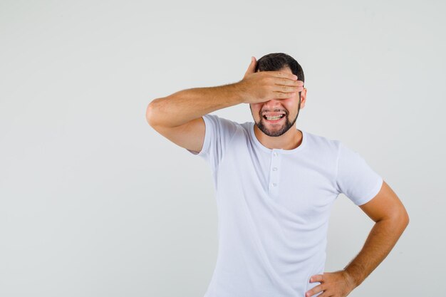 Jeune homme tenant les yeux de la main en t-shirt et l'air joyeux. vue de face.