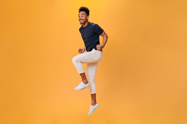 Photo gratuite jeune homme en tenue élégante sautant joyeusement sur fond orange portrait de mec en pantalon blanc se déplaçant sur fond isolé