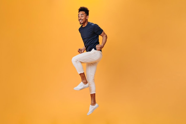 Jeune homme en tenue élégante sautant joyeusement sur fond orange Portrait de mec en pantalon blanc se déplaçant sur fond isolé