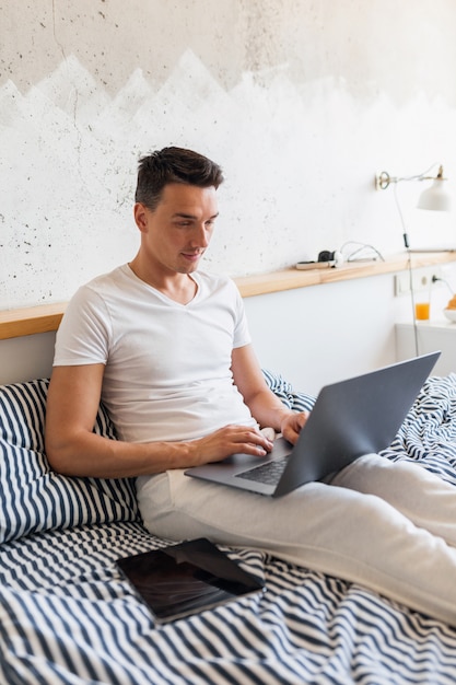 Jeune homme en tenue de pyjama décontracté assis dans son lit le matin travaillant sur ordinateur portable, pigiste occupé à la maison
