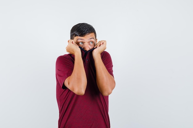 Jeune homme tirant le col sur le visage en t-shirt et ayant l'air effrayé, vue de face.