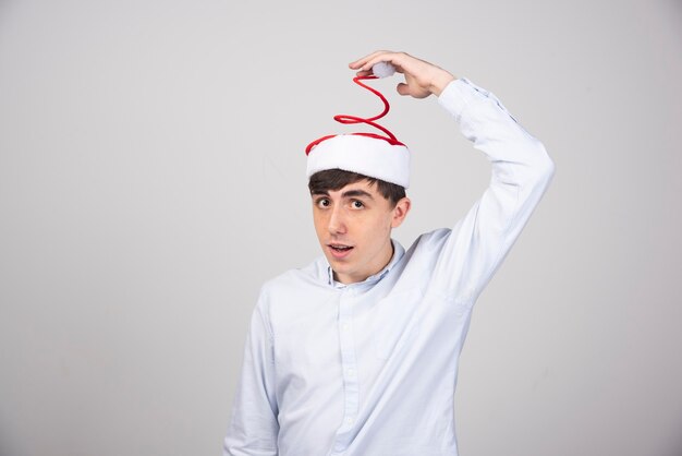 Jeune homme touchant le bonnet de Noel rouge sur le mur gris.