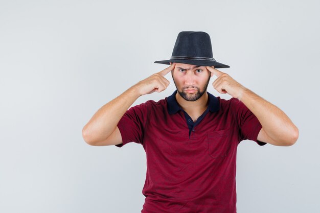 Jeune homme touchant sa tête en t-shirt, chapeau et à la grave, vue de face.