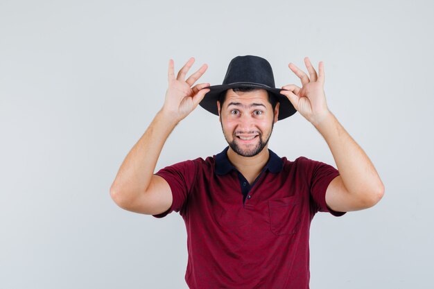 Jeune homme touchant son chapeau avec les doigts en t-shirt, chapeau et à l'optimiste. vue de face.