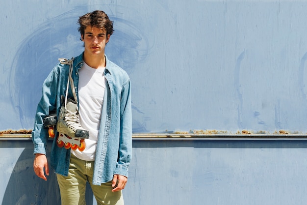 Jeune homme transportant des patins à roulettes debout sur fond bleu en regardant la caméra