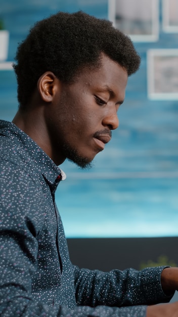 Jeune homme travaillant à domicile, prenant des notes sur le bloc-notes tout en travaillant à distance sur un PC depuis le salon