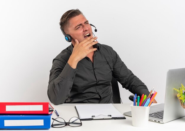 Jeune homme travailleur de bureau blonde agacé sur les écouteurs est assis au bureau avec des outils de bureau à l'aide d'un ordinateur portable crie à quelqu'un regardant à côté isolé sur fond blanc avec espace de copie