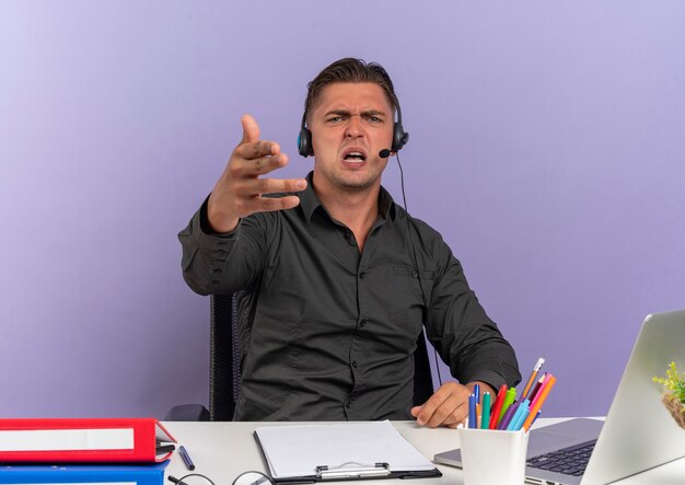 Jeune homme travailleur de bureau blonde agacé sur les écouteurs est assis au bureau avec des outils de bureau à l'aide d'un ordinateur portable regarde et pointe vers la caméra isolée sur fond violet avec espace de copie