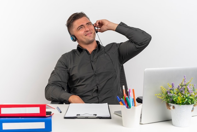 Photo gratuite jeune homme travailleur de bureau blonde agacé sur les écouteurs est assis au bureau avec des outils de bureau à l'aide d'un ordinateur portable tient des écouteurs essayant d'entendre isolé sur fond blanc avec espace de copie
