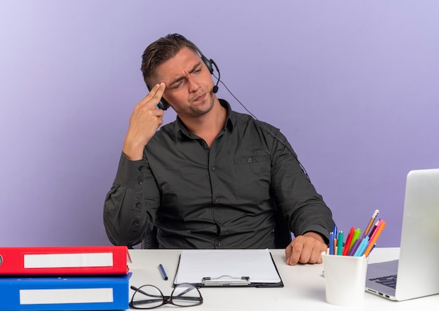 Jeune homme travailleur de bureau blonde agacé sur les écouteurs est assis au bureau avec des outils de bureau à la recherche d'un ordinateur portable met la main sur la tête isolé sur fond violet avec espace de copie