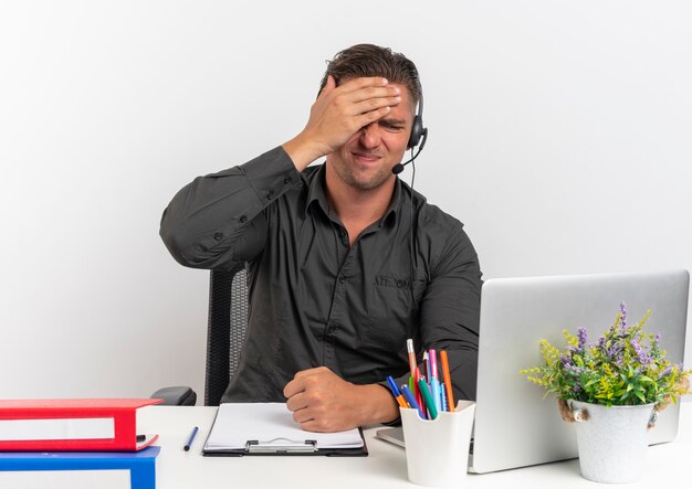 Jeune homme travailleur de bureau blonde bouleversé sur les écouteurs est assis au bureau avec des outils de bureau à l'aide d'un ordinateur portable met la main sur la tête isolé sur fond blanc avec espace de copie