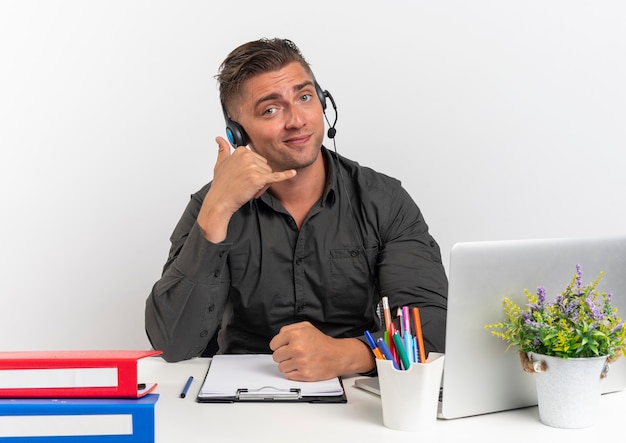 Jeune homme de travailleur de bureau blonde confiant sur le casque est assis au bureau avec des outils de bureau à l'aide de téléphone portable gestes isolé sur fond blanc avec espace de copie