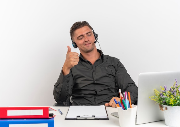 Jeune homme de travailleur de bureau blonde confiante sur les écouteurs est assis au bureau avec des outils de bureau regardant ordinateur portable Thumbs up