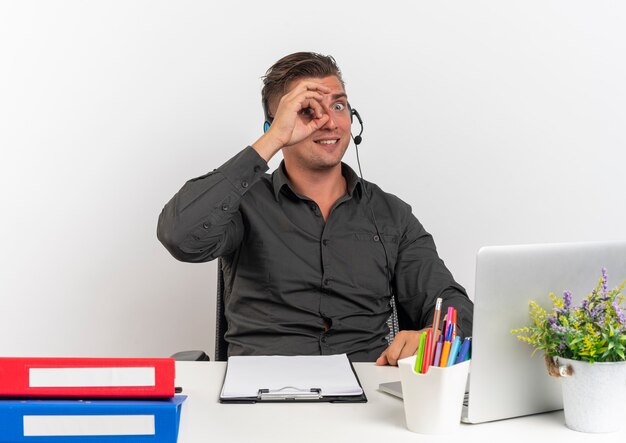 Jeune homme travailleur de bureau blonde surpris sur le casque est assis au bureau avec des outils de bureau à l'aide d'un ordinateur portable regarde à travers les doigts isolés sur fond blanc avec espace de copie