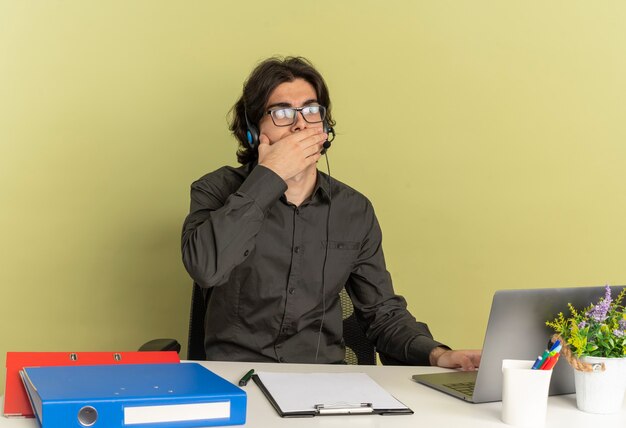 Jeune homme de travailleur de bureau choqué sur les écouteurs dans des lunettes optiques est assis au bureau avec des outils de bureau à l'aide et en regardant un ordinateur portable met la main sur la bouche isolé sur fond vert avec espace de copie