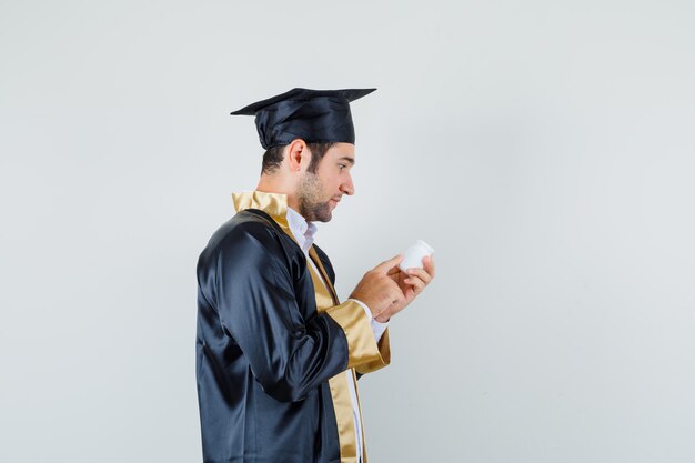 Jeune homme en uniforme de diplômé lisant des informations sur une bouteille de pilules et à la recherche de curiosité.