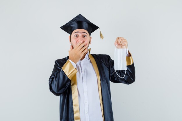 Jeune homme en uniforme de diplômé tenant un masque médical et regardant surpris, vue de face.