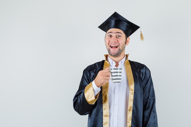 Jeune homme en uniforme de diplômé tenant une tasse de café et regardant joyeux, vue de face.