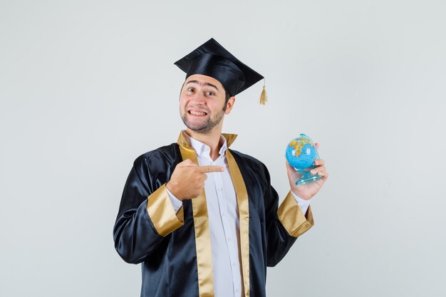 Jeune homme en uniforme d'études supérieures pointant sur le globe de l'école et à la joyeuse vue de face.