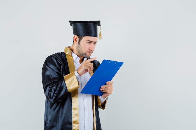 Jeune homme en uniforme d'études supérieures à la recherche de notes sur le presse-papiers, vue de face.