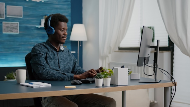 Jeune homme utilisant des écouteurs pour écouter de la musique tout en travaillant du bureau à domicile sur l'ordinateur