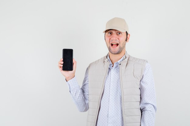 Jeune homme en veste beige et casquette montrant le téléphone et à la surprise, vue de face.