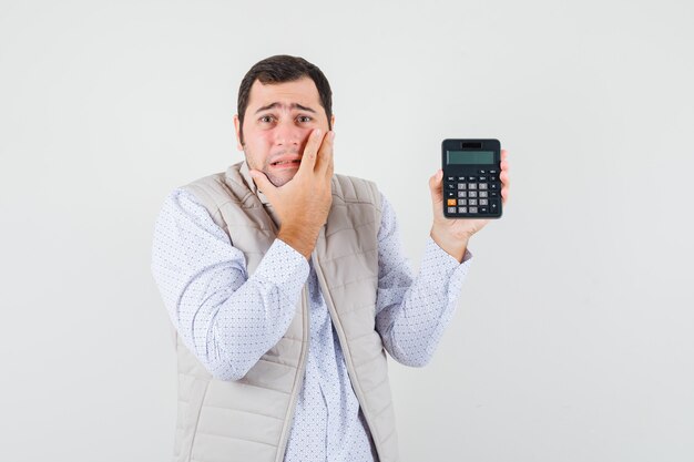 Jeune homme en veste beige et casquette tenant la calculatrice dans une main tout en couvrant une partie du visage avec la main et à la surprise, vue de face.