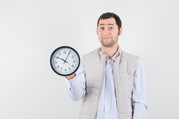 Jeune homme en veste beige et casquette tenant l'horloge en un et à l'optimiste, vue de face.