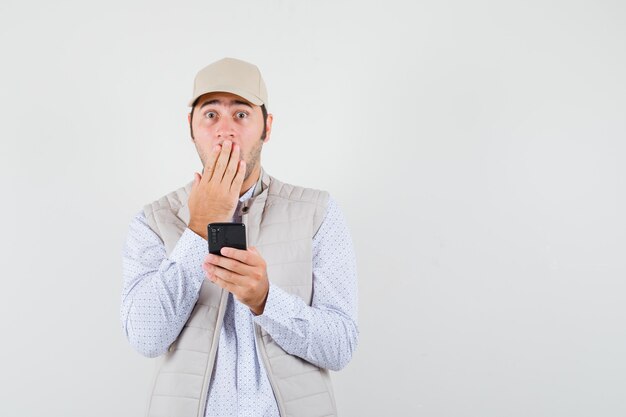Jeune homme en veste beige et casquette tenant le téléphone portable à la main et couvrant la bouche avec la main et à la vue de face, surpris.