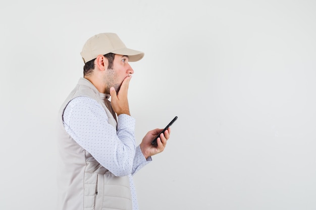 Jeune homme en veste beige et casquette tenant le téléphone portable à la main et couvrant la bouche avec la main et à la vue de face, surpris.