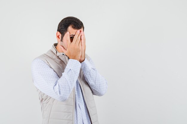 Jeune homme en veste beige couvrant le visage avec les mains et regardant à travers les doigts et à l'optimiste, vue de face.