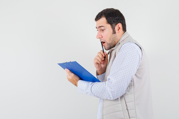 Jeune homme en veste beige mordant le stylo tout en regardant le cahier et à la surprise, vue de face.