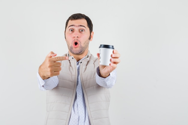 Jeune homme en veste beige pointant vers une tasse de café à emporter avec l'index et à la surprise, vue de face.