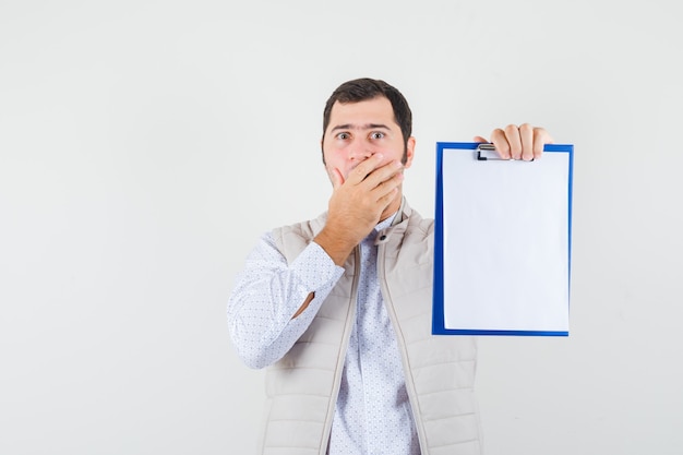 Photo gratuite jeune homme en veste beige tenant le cahier et couvrant la bouche avec la main et l'air surpris, vue de face.