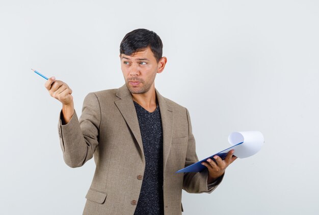 Jeune homme en veste brun grisâtre pointant vers l'extérieur tout en tenant le cahier et à la vue de face, concentré.