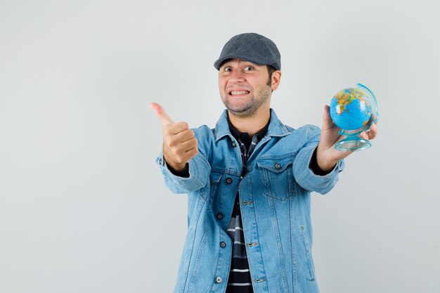 Jeune homme en veste, cap tenant le globe tout en montrant le pouce vers le haut et à la joie