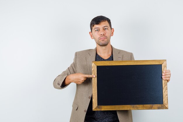 Jeune homme en veste marron grisâtre, chemise noire pointant vers le carton et à la vue de face, prêt.