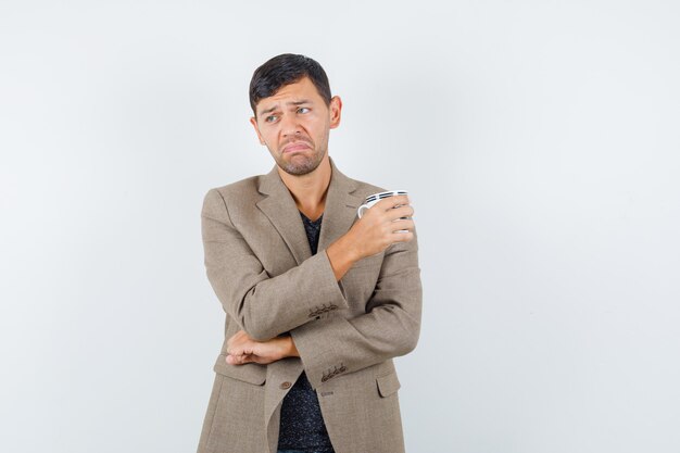 Jeune homme en veste marron grisâtre tenant la tasse et à la vue de face, mécontent.