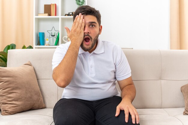 Jeune homme en vêtements décontractés à l'air confus et surpris couvrant les yeux avec la main assis sur un canapé dans un salon lumineux