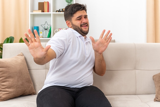 Jeune homme en vêtements décontractés à l'air inquiet en faisant un geste de défense avec les mains assis sur un canapé dans un salon lumineux